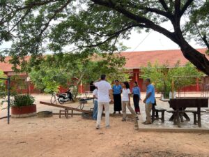 Eine Gruppe von Erwachsenen und Schülern unterhält sich unter einem großen Baum auf einem Schulhof, im Hintergrund sind rote Gebäude zu sehen.